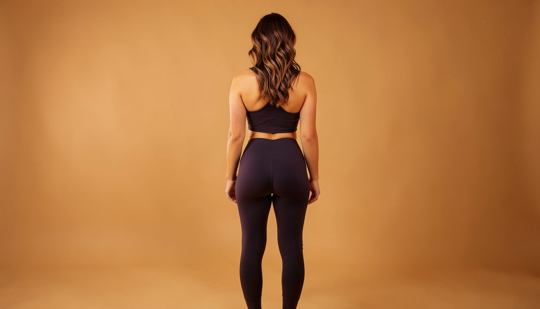 Rear view of a woman in fitted leggings highlighting a lifted silhouette, warm studio lighting